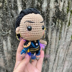 K&ouml;nnte beinhalten: H&auml;kelpuppe mit schwarzen Haaren, brauner Haut und blau-violetten Kleidungsst&uuml;cken. Die Puppe tr&auml;gt goldene Ohrringe und hat schwarze Augen mit gelben Pupillen.