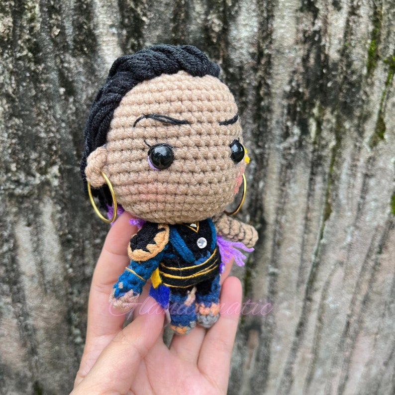 K&ouml;nnte beinhalten: H&auml;kelpuppe mit schwarzen Haaren, brauner Haut und blau-violetten Kleidungsst&uuml;cken. Die Puppe tr&auml;gt goldene Ohrringe und hat schwarze Augen mit gelben Pupillen.