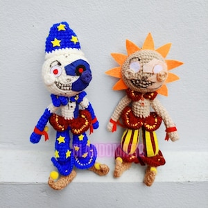 May include: Two crocheted plush toys, one with a blue and white hat and the other with a yellow and orange sun-shaped head. Both toys have red and yellow clothing and a creepy smile.