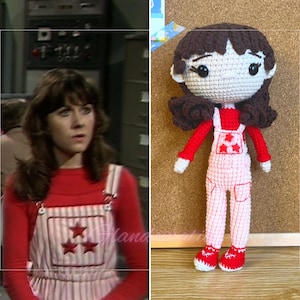 Puede incluir: Una muñeca de crochet que lleva una camisa roja y un mono rosa con estrellas blancas. La muñeca tiene el pelo castaño y una cara blanca con ojos negros.