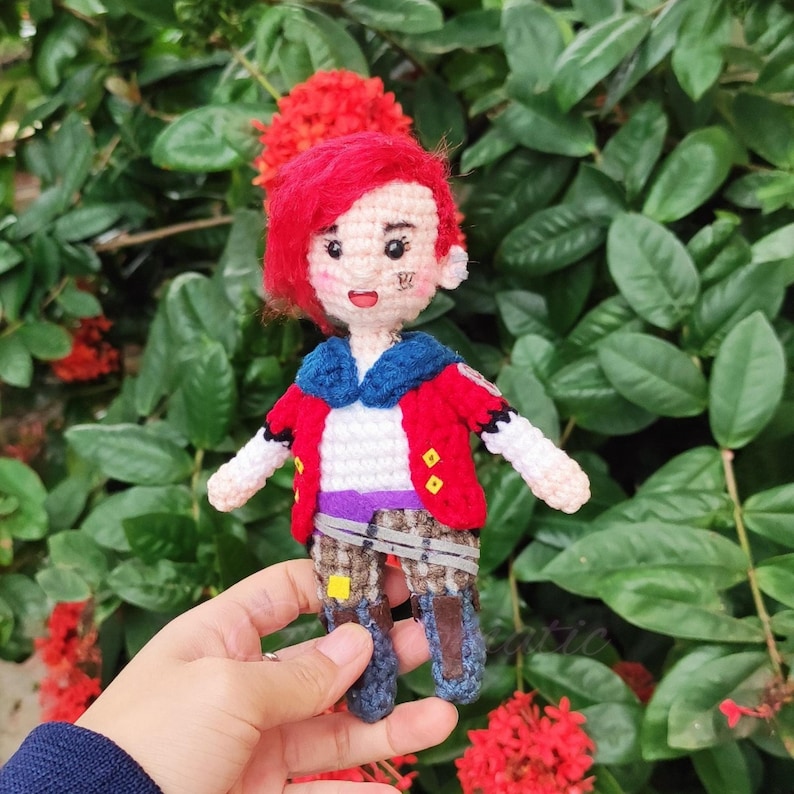 May include: A crocheted doll with red hair, a red and blue jacket, and a purple belt. The doll is wearing brown pants and black shoes. The doll is holding a small red heart in its hand.