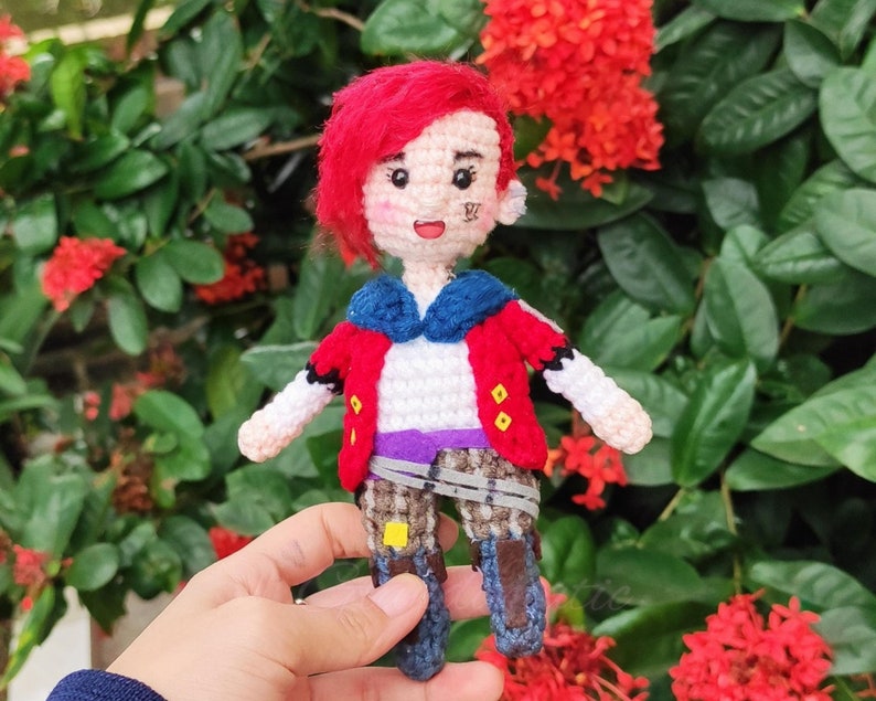 May include: A crocheted doll with red hair, a red jacket, a blue shirt, and a purple belt. The doll is wearing brown boots and has a yellow and black design on its legs. The doll is holding a small, yellow object in its hand.