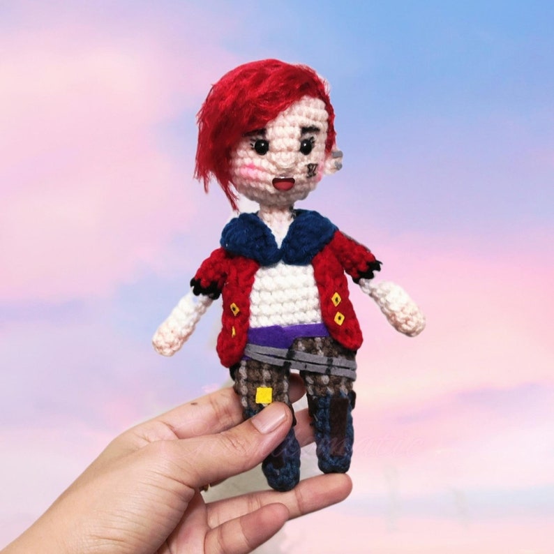 May include: A crocheted doll with red hair, a red and blue jacket, and purple and brown pants. The doll has a painted face with a black eye patch.