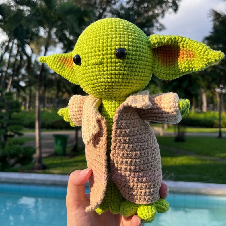 May include: A handmade green crocheted doll with large ears and black eyes, wearing a tan and brown coat. The doll is a character from a popular science fiction franchise. The doll is approximately 10 inches tall.
