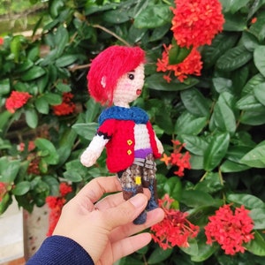 May include: A crocheted doll with red hair, a red and blue jacket, and a blue and white skirt. The doll is holding a small, blue and white bag. The doll is standing on a hand.