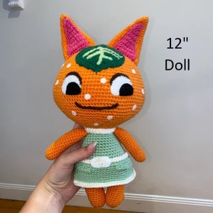 Pu&ograve; includere: Una bambola di 30,5 cm realizzata a uncinetto, raffigurante un gatto arancione con una foglia verde sulla testa. La bambola indossa un vestito verde con una cintura bianca.