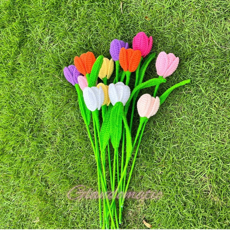 May include: A bouquet of handmade crochet tulips in various colors, including orange, purple, pink, white, and yellow. The flowers have green stems and leaves, arranged against a backdrop of green grass.
