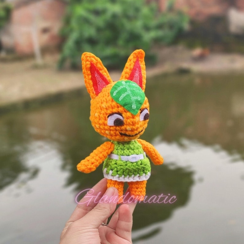 Pu&ograve; includere: Un giocattolo di peluche all'uncinetto arancione e verde con una foglia sulla testa. Il giocattolo &egrave; tenuto da una mano.