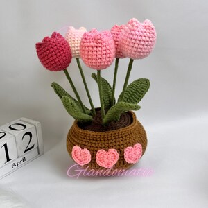 Peut inclure: Un bouquet de tulipes crochet&eacute;es roses et rouges dans un pot brun avec des accents de c&oelig;ur roses. Le pot est pos&eacute; sur une surface blanche avec un calendrier en bois &agrave; blocs qui indique "Avril 12".