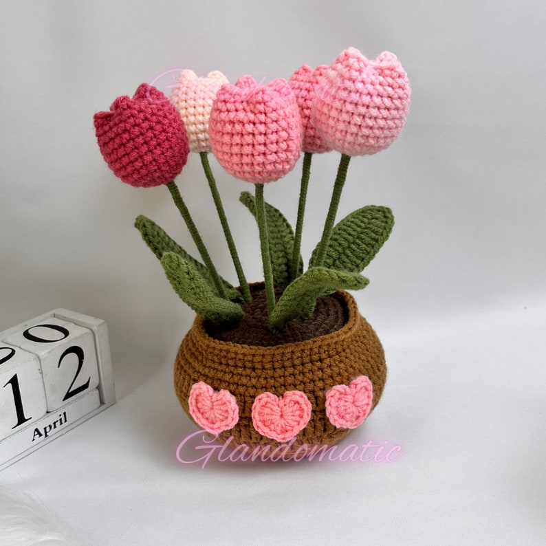 Peut inclure: Un bouquet de tulipes crochet&eacute;es roses et rouges dans un pot brun avec des accents de c&oelig;ur roses. Le pot est pos&eacute; sur une surface blanche avec un calendrier en bois &agrave; blocs qui indique "Avril 12".