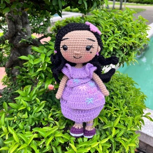 May include: A crocheted doll with long black hair, wearing a purple dress with floral embroidery and purple shoes. The doll is sitting on a bed of green leaves.