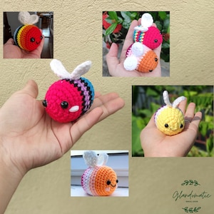 May include: Five crocheted bee plushies in various colors, including pink, yellow, orange, and blue. Each bee has white wings and black eyes. The bees are arranged in different positions, some with their wings spread out and others with their wings folded.