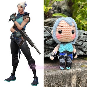 Pu&ograve; includere: Una bambola amigurumi all'uncinetto di un personaggio femminile con capelli blu, un top blu e bianco e pantaloni grigi. La bambola tiene una pistola nella mano destra. La bambola &egrave; seduta su uno scoglio.