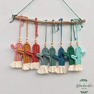Könnte beinhalten: Ein Set aus acht bunten Makramee-Kaktus-Wandbehängen mit Holzperlen und Quasten. Die Kakteen sind in den Farben Rosa, Orange, Rot, Grün und Blau gehalten. Die Behänge sind an einem natürlichen Ast befestigt.