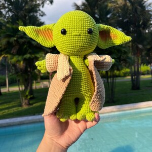 May include: A handmade crochet doll of a character with large ears and black eyes. The doll is primarily green with a tan coat and is held in a hand. The doll is a Star Wars character.