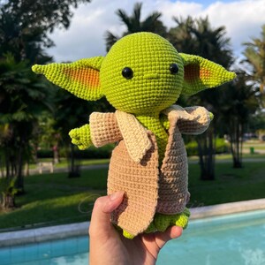 May include: A crocheted doll of a character with large ears, green skin, and black eyes. The doll wears a tan coat and a brown robe. The doll is held in front of a blurred background of trees and a water feature.