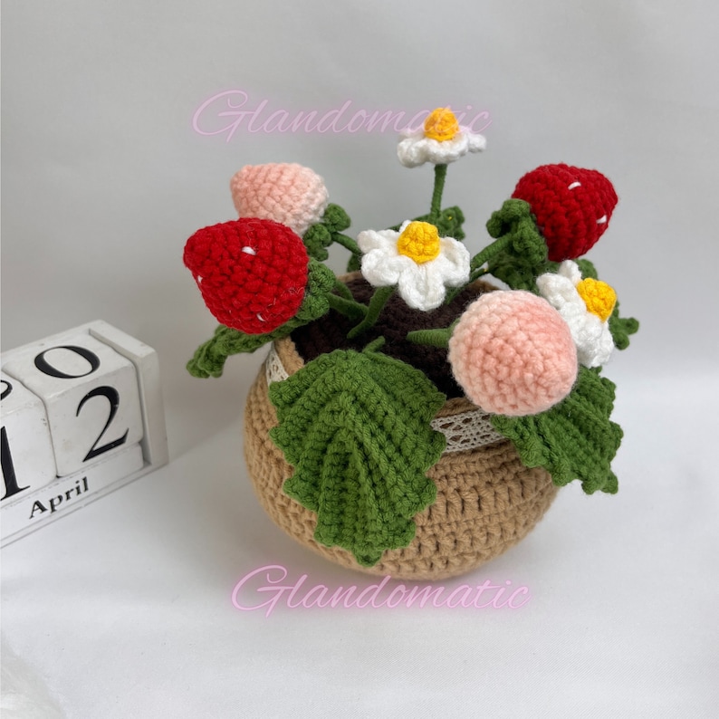 Peut inclure: Une plante de fraises en crochet dans un pot brun avec une bordure en dentelle blanche. La plante a des fraises roses et rouges, des fleurs blanches avec des centres jaunes et des feuilles vertes.