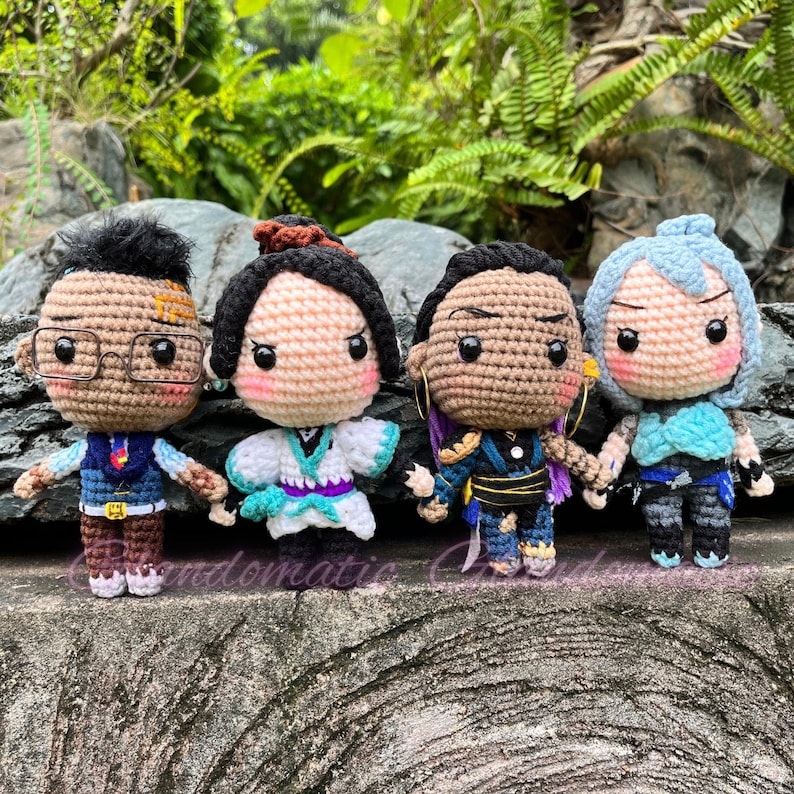 K&ouml;nnte beinhalten: Vier geh&auml;kelte Pl&uuml;schfiguren, jede mit einzigartigen Gesichtsausdr&uuml;cken und Kleidung. Die Figuren stehen auf einer strukturierten Oberfl&auml;che. Die Figuren tragen eine Brille, ein wei&szlig;-blaues Kimono, ein lila-schwarzes Outfit und ein blau-wei&szlig;es Outfit.