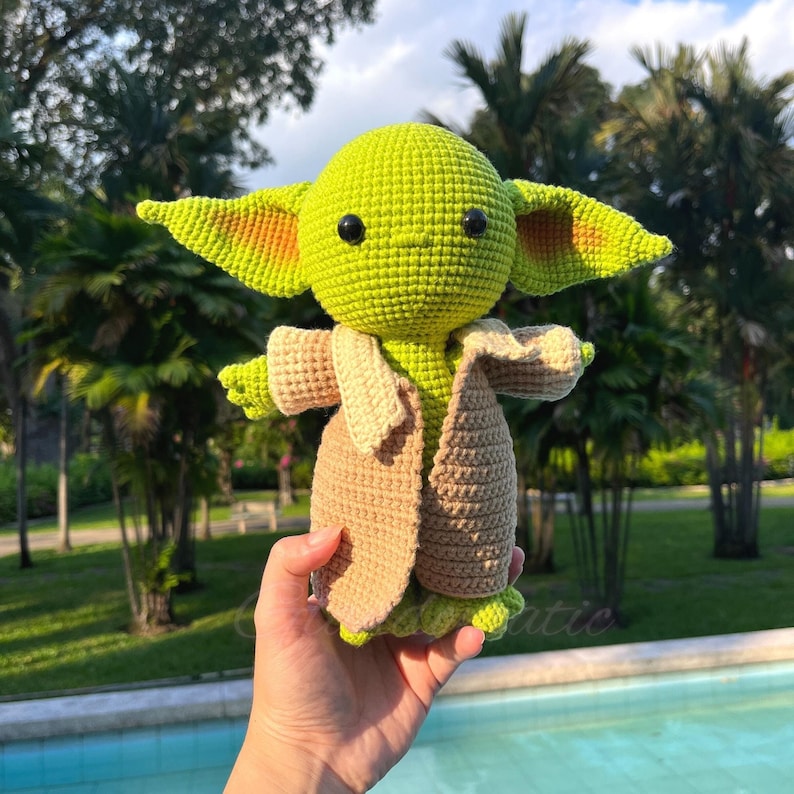 May include: A handmade crochet doll of a character with large ears, green skin, and black eyes. The doll wears a tan coat and a light green outfit. The doll is held in front of a background of green trees and a pool.