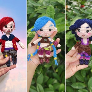 May include: Three crocheted figures, each with a different hairstyle and outfit. The figure on the left has red hair and a red jacket, the middle figure has blue hair and a purple top, and the figure on the right has blue hair and a purple top.