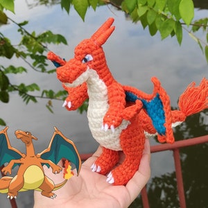 Op de afbeelding: Een gehaakt oranje en blauw Charizard Pokémon knuffel. Het speelgoed heeft een witte buik en een lange staart. Het speelgoed wordt in de hand van een persoon gehouden.