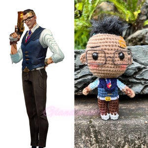 Pu&ograve; includere: Una bambola amigurumi all'uncinetto con pelle marrone, capelli marroni e un gilet blu. La bambola indossa gli occhiali e una camicia blu chiaro sotto il gilet. La bambola tiene un piccolo cuore rosso nella mano.
