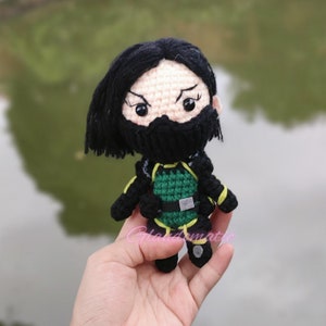 May include: Crocheted doll of a character with black hair, a black face covering, and a green and black outfit. The doll has a stern expression with black eyes and eyebrows. The doll is held in a hand against a blurred green background.
