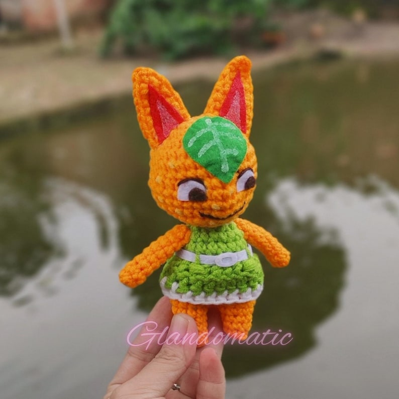 Pu&ograve; includere: Un giocattolo di peluche all'uncinetto arancione e verde con una foglia sulla testa. Il giocattolo indossa un vestito verde con una cintura bianca.