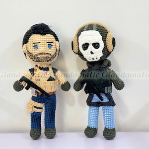 May include: Two handmade crochet dolls. One doll has a black beard, blue eyes, and a tan vest with a black gun. The other doll has a skull mask and a green and black outfit. Both dolls are standing.