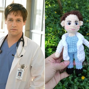 May include: A crochet doll of a doctor wearing a white coat, blue scrubs, and black shoes. The doll has brown hair and a stethoscope around its neck. The doll is held in a hand against a background of green foliage.