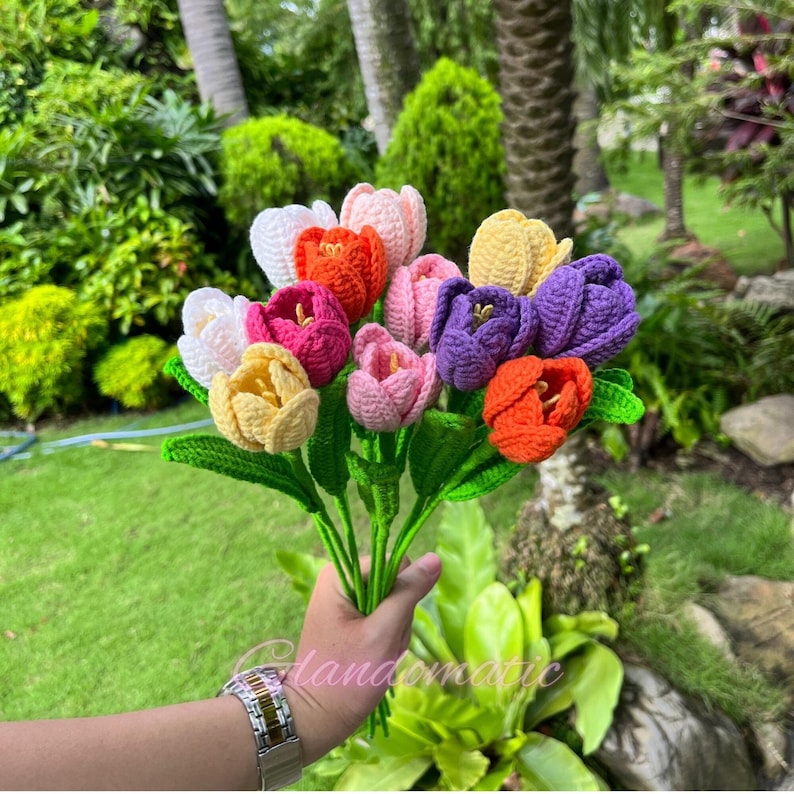 May include: A bouquet of handmade crochet tulips in various colors, including white, orange, pink, purple, and yellow. The flowers have green stems and leaves, held together. The background features lush green foliage.