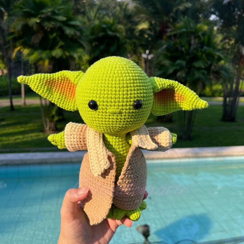 May include: A handmade green crochet figure with large ears, black eyes, and a tan robe. The figure is a character from a popular science fiction franchise. The figure is approximately 6 inches tall.