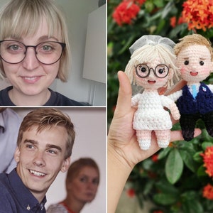 May include: A crocheted bride and groom doll set. The bride is wearing a white dress with a veil and glasses. The groom is wearing a blue suit with a white shirt and a blue tie. The dolls are holding hands and are standing on a hand.