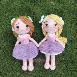 May include: Two crocheted dolls with long hair, wearing purple dresses and white flower headbands. The dolls are sitting on a bed of green grass.
