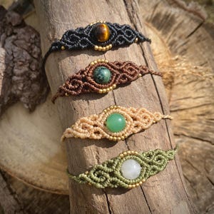 Könnte beinhalten: Vier Makramee-Armbänder mit Edelstein-Akzenten werden auf einer Holzoberfläche präsentiert. Die Armbänder haben schwarzes, braunes, beiges und grünes Makramee mit Tigerauge-, grünen und weißen Edelsteinen. Jedes Armband hat goldfarbene Perlen.
