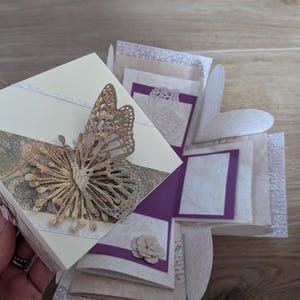 Könnte beinhalten: Eine handgefertigte Explosionsbox mit Schmetterlingsverzierungen. Die Box ist cremefarben mit einem dekorativen gemusterten Band und einem goldenen Schmetterling. Im Inneren befinden sich lila und weiße Dekorationselemente. Die Box ist geöffnet und zeigt mehrere Schichten.