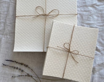 May include: Two stacks of off-white, textured paper tied with twine bows. The paper has a subtle, repeating pattern. A few sprigs of dried lavender are placed near the paper stacks on a light gray linen surface.
