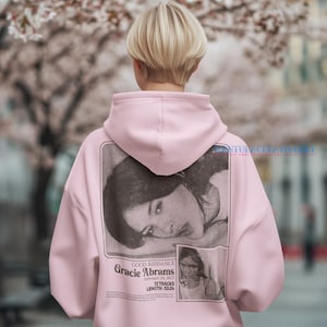 May include: A pink hoodie with a black and white photo of a woman on the back. The photo has the text "GOOD RIDDANCE Gracie Abrams February 24, 2023 12 TRACKS LENGTH: 52:24" printed below it.