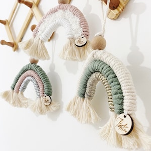 May include: Four macrame rainbow decorations in pastel shades, suspended against a white wall. Each rainbow has a unique colour scheme, with a wooden bead and tassel. A wooden tag with text is attached to each rainbow.