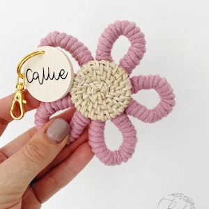 May include: A macrame flower keyring in a daisy design. The flower features dusty rose petals and a woven natural centre. A wooden tag with the name "Callie" is attached with a gold-coloured clasp.