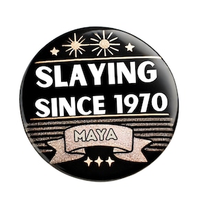 Puede incluir: Botón negro y dorado con el texto "SLAYING SINCE 1970" y el nombre "MAYA" en una pancarta.
