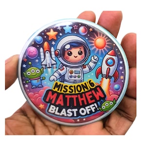 Puede incluir: Un botón redondo con un diseño colorido de temática espacial. Presenta un astronauta de dibujos animados con un cohete, estrellas, planetas y el texto "Misión 6 Matthew Blast Off!"