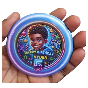 May include: A blue and purple round button with a neon design. The button features a cartoon illustration of a young boy with a happy expression. The text "Happy Birthday Jayden 5" is written in a neon font.