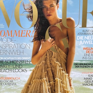 Può includere: Una copertina della rivista Vogue con una donna in un abito beige a balze. Il titolo della rivista è dorato, con la parola "SOMMER!" in arancione. Lo sfondo mostra una scena di spiaggia. Il testo include "MODE-INSPIRATION" e "BEACH-LOOKS."