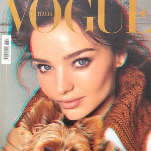 Könnte beinhalten: Vogue Italia Magazin-Cover mit einer Frau und einem kleinen Hund. Der Titel des Magazins ist in Goldbuchstaben gehalten. Der Cover hat einen 3D-Effekt. Der Text "VISION OF FEMININITY" befindet sich unten.