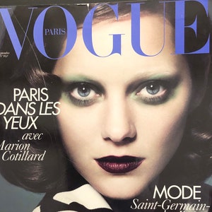 以下が含まれることがあります： マリオン・コティヤールを表紙に迎えたヴォーグ誌。緑のアイシャドウと濃い赤のリップをつけたマリオン・コティヤール。表紙のテキストは「パリ・ダン・レ・ジュー」と「モード・サンジェルマン・デ・プレ、セ・ノワール」で、ジュリエット・グレコが自信を持って語っている。