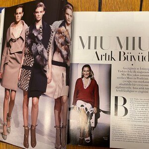May include: A magazine spread showcasing a fashion editorial featuring three models in stylish winter attire. The models are wearing coats, dresses, and boots. The magazine spread also includes a photograph of Miuccia Prada, the designer of the Miu Miu brand, and text about the brand's expansion and growth.