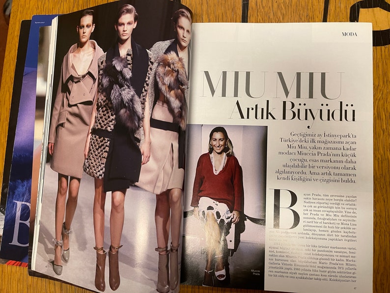 May include: A magazine spread showcasing a fashion editorial featuring three models in stylish winter attire. The models are wearing coats, dresses, and boots. The magazine spread also includes a photograph of Miuccia Prada, the designer of the Miu Miu brand, and text about the brand's expansion and growth.