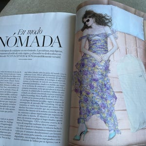 Puede incluir: Una p&aacute;gina de revista de moda con una mujer con un vestido floral y tacones azules, acostada en una cama. El vestido tiene un dise&ntilde;o en capas con flores moradas, amarillas y blancas. El texto "En modo NOMADA" es visible.