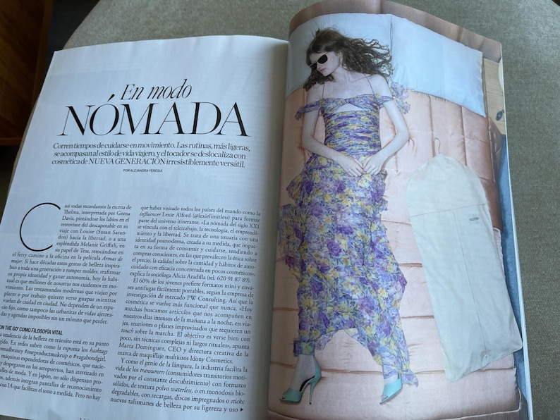 Puede incluir: Una p&aacute;gina de revista de moda con una mujer con un vestido floral y tacones azules, acostada en una cama. El vestido tiene un dise&ntilde;o en capas con flores moradas, amarillas y blancas. El texto "En modo NOMADA" es visible.